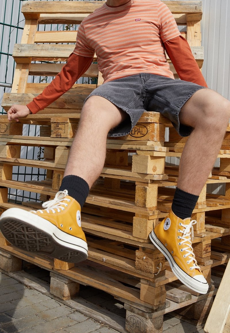 Homme portant une chemise rayée orange et un short gris assis sur des palettes en bois empilées, jambes étendues, baskets montantes jaunes visibles.