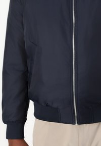 Marineblå bomberjakke med frontglidelås, ribbede mansjetter og hem. Glatt tekstur med minimal detaljering og et moderne, tettsittende design.