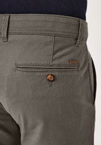 Graue Baumwollhosen mit einer Gesäßtasche, die mit einem braunen Knopf gesichert ist, und einem kleinen Markenschild nahe dem Bund. Glatte Textur.