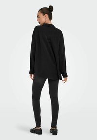 Camisa negra de botones con mangas largas, botones en contraste y un corte relajado, combinada con jeans negros ajustados y zapatos planos.