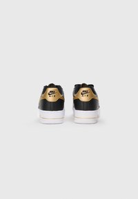 Sapatilhas Nike Air em preto e dourado com um cabedal de couro liso, sola de borracha branca e detalhes dourados no calcanhar e na lateral.