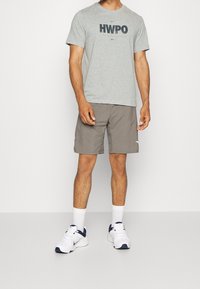 Man som bär en grå HWPO-t-shirt, taupe shorts, vita strumpor och vita samt marinblå Nike-sneakers och står mot en enkel vit bakgrund.
