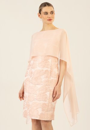 Frau trägt ein ärmelloses, knielanges, hellrosa Blumenkleid mit einem transparenten, asymmetrischen Umhang-Overlay und steht vor einem schlichten Hintergrund.