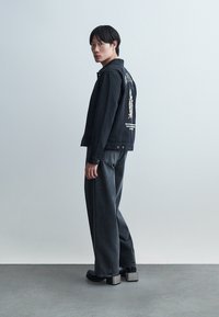 Tânăr purtând o jachetă neagră din denim cu imprimeu grafic pe spate, blugi largi de culoare închisă și pantofi cu platformă, stând pe lateral.