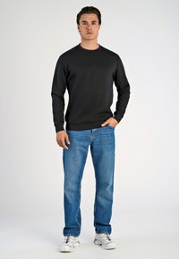 Felpa nera con scollo rotondo, abbinata a jeans blue e sneaker grigie. Il modello posa con le mani in tasca su uno sfondo neutro.