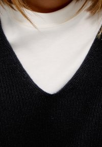 Zwarte gebreide vest met een diepe V-halslaag over een witte coltrui. De textuur van de stof is zichtbaar, wat het geweven patroon benadrukt.