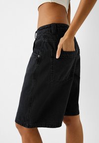 Mustat denimshortsit, joissa on rento istuvuus, sivutaskut ja näkyvät ompelutahdet. Materiaalilla on sileä, hieman teksturoitu ulkonäkö.