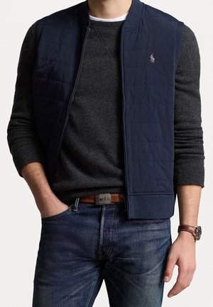 Homme portant une veste matelassée bleu marine sur un pull gris foncé, un jean bleu, une ceinture marron et une montre-bracelet avec un bracelet en cuir marron.