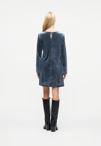 Γυναίκα με μακριά ξανθά μαλλιά που φοράει μπλε βελούδινο φόρεμα με μακριά μανίκια και μαύρες μπότες μέχρι το γόνατο, στέκεται προς τα πίσω σε λευκό φόντο.