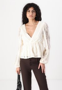 Peplum-bluse i gjennomsiktig kremfarge med blonder i blomsterdesign, dyp V-hals og lange, luftige ermer; kombinert med brune tettsittende bukser.