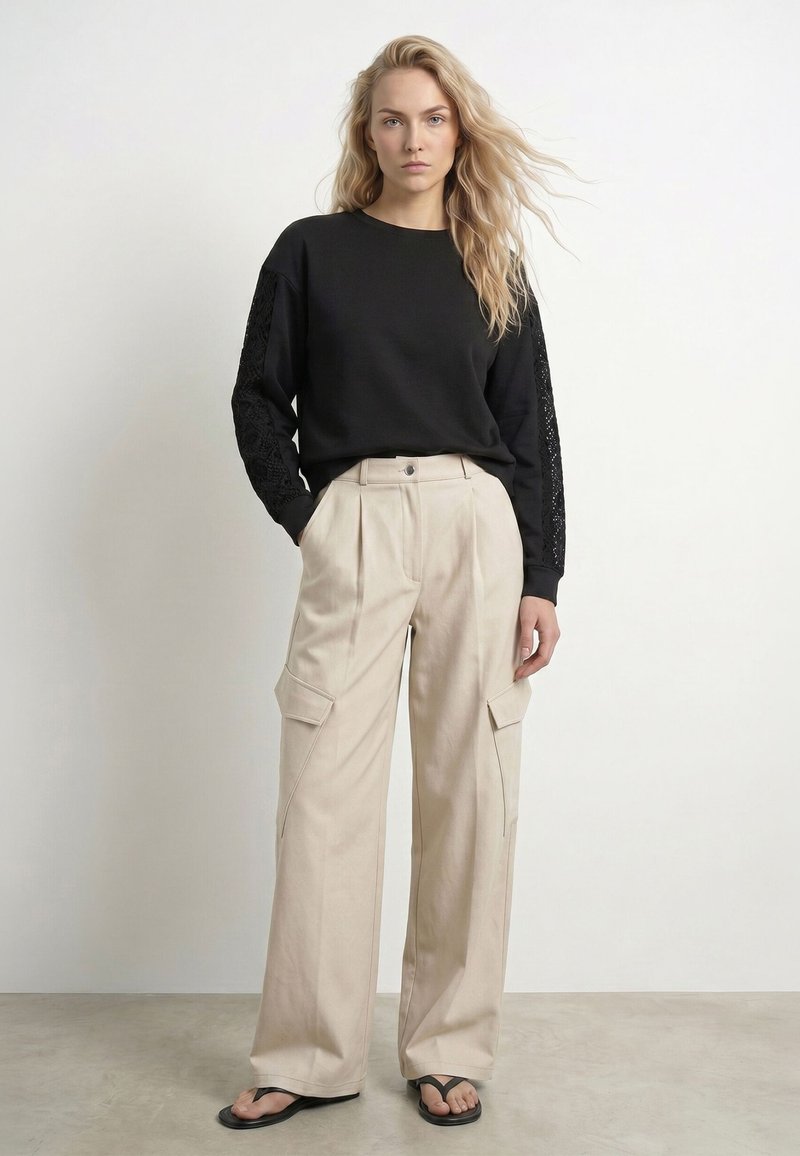 Femme aux cheveux blonds lâchés portant un haut noir à manches longues avec des manches en dentelle, un pantalon large beige avec des poches et des sandales noires, debout.