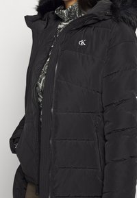 Chaqueta acolchada negra de Calvin Klein con capucha adornada con pelo, textura acolchada, cierre delantero con cremallera y detalle del logo en el pecho.