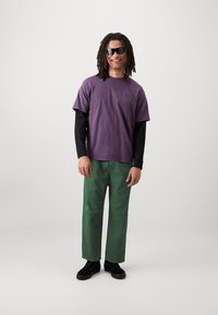 Paarse korte mouwen t-shirt over een zwarte lange mouwen shirt, groene broek en zwarte sneakers. Model draagt grote zwarte zonnebril.