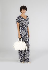 Yksivärinen kukkakuvioinen haalari tummansinisessä ja valkoisessa värissä, lyhyillä hihoilla. Pitelee tekstuuriltaan valkoista käsilaukkua, jossa on tupsuja. Vaalean väriset kengät.