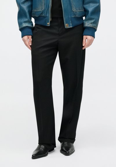 Pantalon noir sur mesure à coupe droite, avec une texture lisse. Porté avec des chaussures en cuir noir et une veste bomber en cuir bleu.