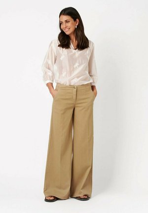 Frau lächelt mit Händen in den Taschen, trägt eine durchsichtige weiße Bluse mit floralen Details, weite beige Hosen und schwarze Sandalen.