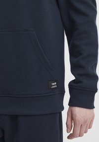 Felpa blu navy con tessuto texturizzato, tasca a marsupio e etichetta nera del brand sull'orlo. Presenta polsini e orlo a costine.