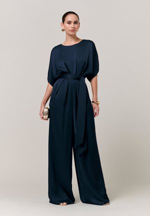 Kvinde i marineblå bredbenet jumpsuit med bælte, der holder en lille guldtaske, iført guld øreringe og armbånd, poserer på neutral baggrund.