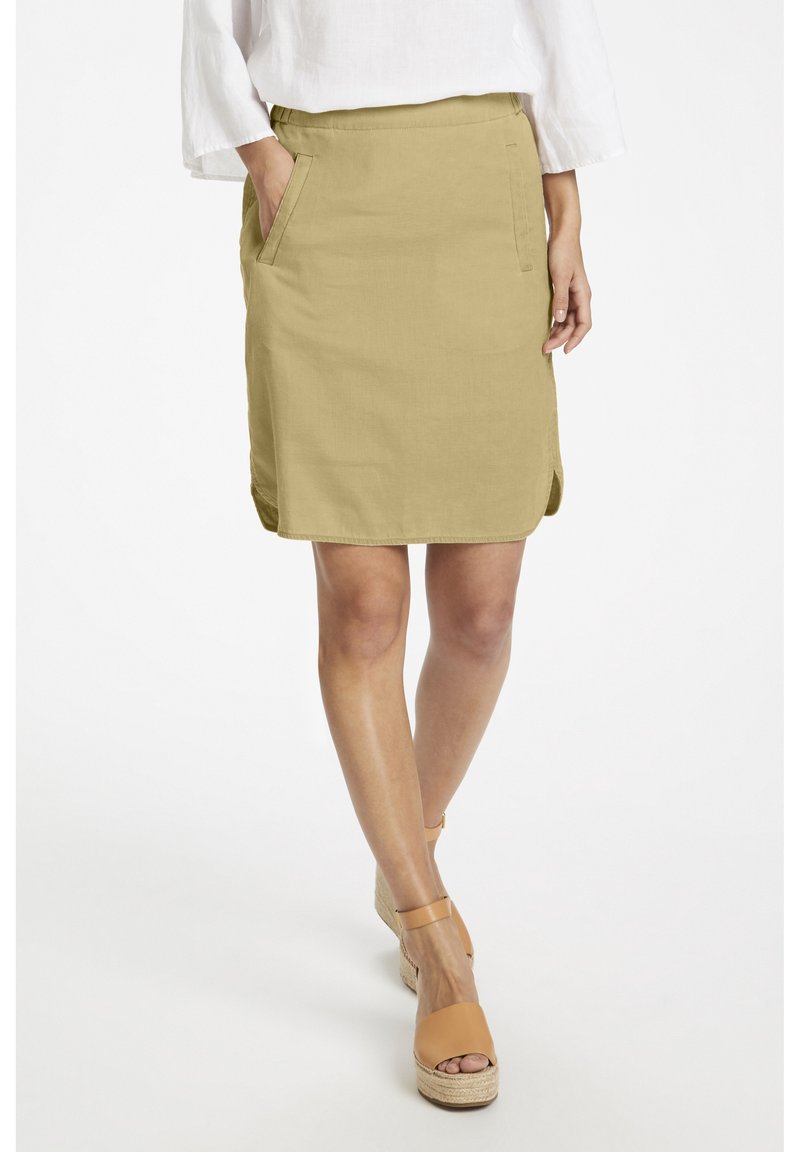 Jupe kaki à coupe droite, avec des poches latérales et un ourlet légèrement incurvé. Portée avec des sandales compensées couleur camel. Texture du tissu lisse.