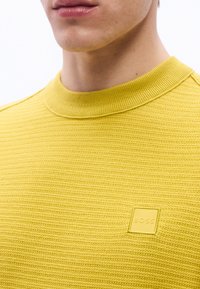 Gros plan sur une personne portant un pull en maille jaune texturée avec un col rond et un petit logo carré "BOSS" sur la poitrine.