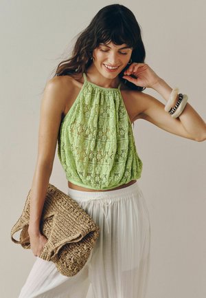 Nő ujjatlan zöld csipkés halter felsőt visel, fehér nadrággal, egy nagy fonott clutchot tart a kezében, és egymásra rakott karkötőket visel az egyik karján.