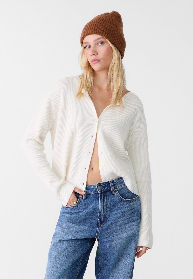 Cardigan bianco a coste con scollo a V e chiusura a bottoni, abbinato a jeans denim blu morbidi. Il modello indossa un berretto di lana marrone.