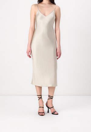 Kobieta ubrana w beżową sukienkę na ramiączkach i czarne sandały na wysokim obcasie z paskami wokół kostki, stojąca na białej podłodze.