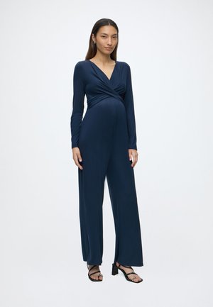 Femme enceinte debout portant une combinaison cache-cœur bleu marine à manches longues et des sandales à talons noires sur un fond blanc uni.