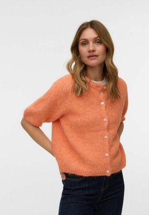 Žena s vlnitými vlasmi po plecia nosí huňatý oranžový krátky sveter na gombíky a tmavé džínsy, stojí s rukami v zadných vreckách.