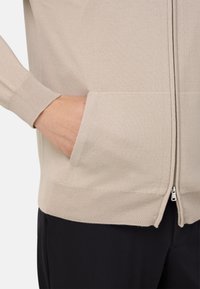 Béžový pletený cardigan so zipsom vpredu, rebrovanými manžetami a bočnými vreckami, charakterizovaný hladkou textúrou a štandardným strihom.