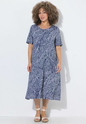 Kobieta z kręconymi włosami, ubrana w niebiesko-białą sukienkę midi w liście oraz beżowe sandały z paskiem wokół kostki, stojąca przy białej ścianie.