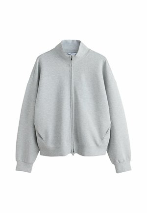 Frau trägt eine hellgraue Zip-up-Jacke und passende Sweatpants mit weißen Sneakers, stehend vor einfachem braunem Hintergrund.
