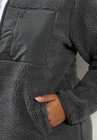 Felpa grigia in pile con corpo testurizzato, tasca sul petto liscia con zip e tasche laterali. Maniche con polsino e bordi a contrasto.