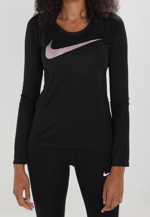 Femme portant un t-shirt à manches longues noir Nike avec un logo swoosh rose et des leggings Nike noirs avec un logo swoosh blanc sur la cuisse.
