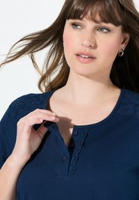 Námořnická modrá bavlněná košile s kulatým výstřihem, dekorativní krajkovou výzdobou na ramenou a třemi fialovými knoflíky na léze.