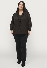 Zwarte blouse met een gerimpelde V-hals, stippenpatroon en lange mouwen. Gecombineerd met zwarte slim-fit broek en hoge enkellaarsjes.