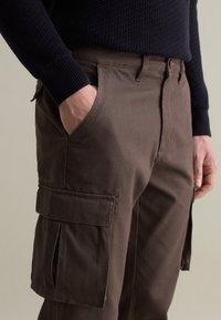 Braune Cargo-Hosen mit Seitentaschen, aus robuster Stoff gefertigt. Mit Gürtelschlaufen und lockerem Schnitt. Tragen mit einem dunkel strukturierten Pullover.