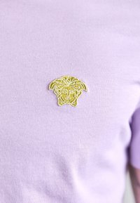 Světlofialová bavlněná košile s žlutým vyšívaným vzorem obličeje s vlasy, umístěným na levé straně hrudi.