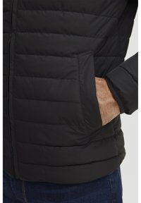 Svart boblejakke med quiltet tekstur, med en sidelomme og elastiske ermer. Materialet fremstår som glatt og vannavvisende.