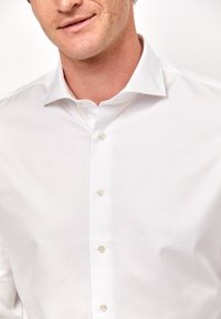 Camicia bianca con colletto classico, caratterizzata da una texture liscia e sei bottoni abbinati lungo la patta anteriore.