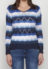 Femme portant un pull en tricot à rayures bleues, blanches et bleu marine avec des motifs géométriques et un jean bleu, debout devant un fond clair uni.