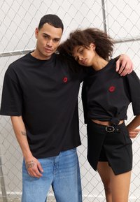Sorte bomulls-t-skjorter med rød leppeprint på brystet, iført med blå denimjeans og et svart omslagskjørt med beltespenne-detalj.