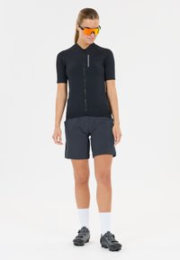 Kvinna i svart kortärmad cykeltröja, mörkgrå shorts, vita strumpor, svarta cykelskor och orange reflekterande sportsolglasögon på vit bakgrund.