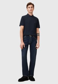 Navyblaues Poloshirt mit kurzen Ärmeln, glatter Textur und dezentem Logo. Kombiniert mit dunkelblauen Jeans im geraden Schnitt und schwarzen Schuhen.