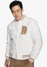 Hvid varsity jakke i uldblanding med en tan "D" patch, broderet tekst samt stripede ribstrikkede manchetter og krave. Knapper til lukning.