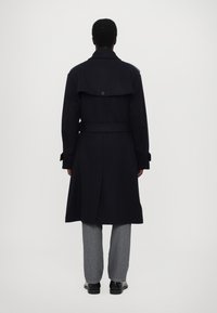 Trench coat navy con vita abbinata, colletto alto e bottone sulla schiena; abbinato a jeans grigi e scarpe nere.