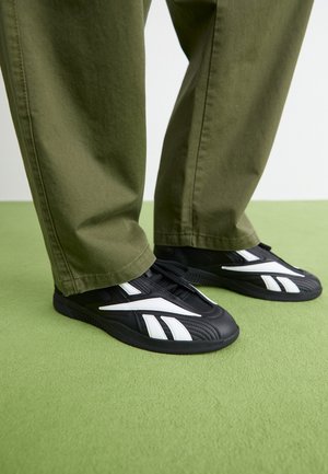 Zapatillas deportivas negras con acentos blancos, que cuentan con una parte superior texturizada y una punta redondeada, combinadas con pantalones verdes oliva.