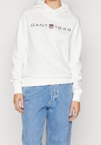 Sudadera blanca con capucha que presenta el texto "GANT 1949" y un emblema en azul oscuro y rojo. Tejido suave, con puños y dobladillo acanalados. Combinada con jeans de color azul claro.