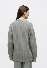 Světlounce šedý svetr z texturovaného úpletu s volným střihem a žebrovanými detaily. Má kulatý výstřih a je nošen přes volné šedé kalhoty.
