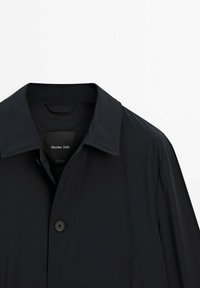 Schwarzer leichter Trenchcoat mit spitzem Kragen, einzelner Knopfverschluss und glatter Textur. Etikett im Inneren des Mantels sichtbar.